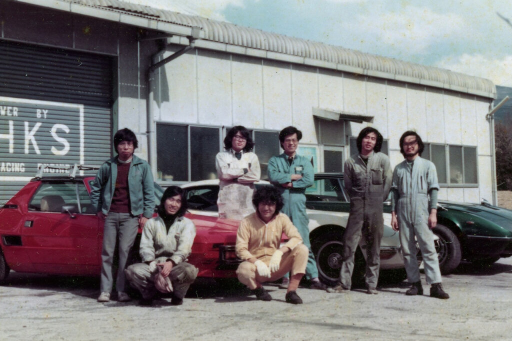 7 people posing in front of cars and the original HKS garage