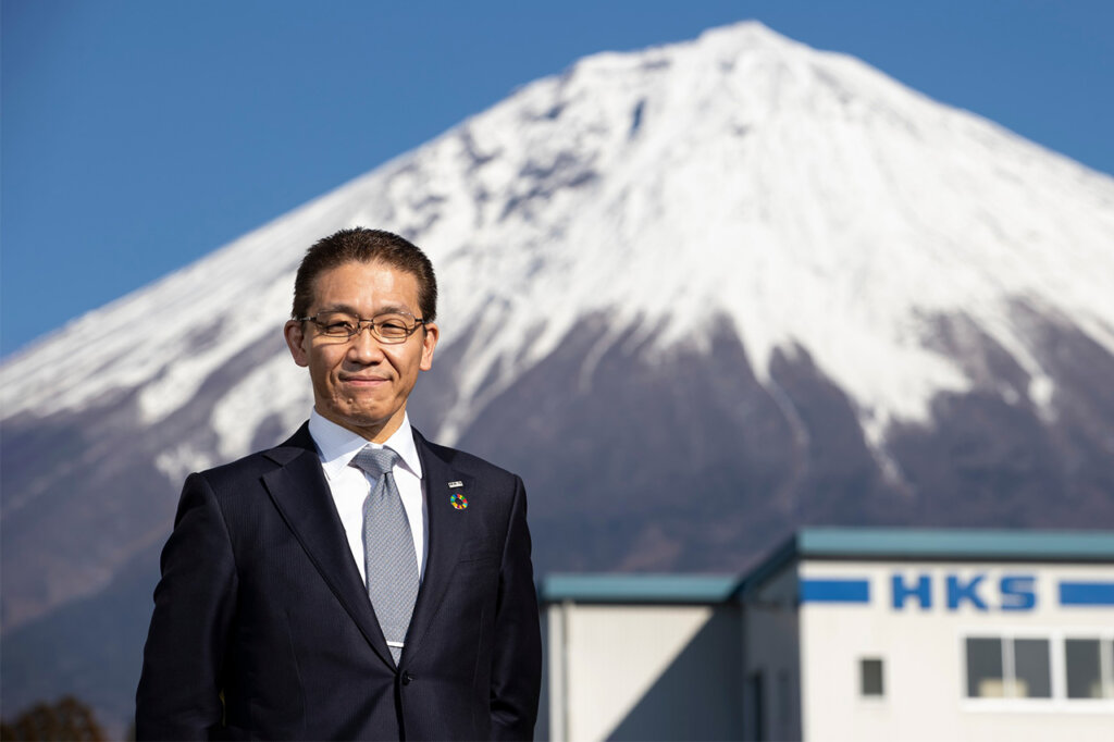 man in a suit next with a building and a snow covered Mt. Fuji behind him