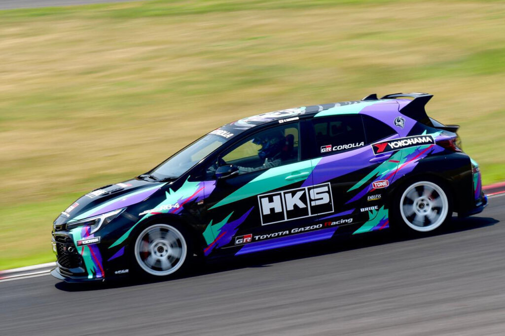 GR Corolla with an HKS Livery driving fast on a racetrack grass in background