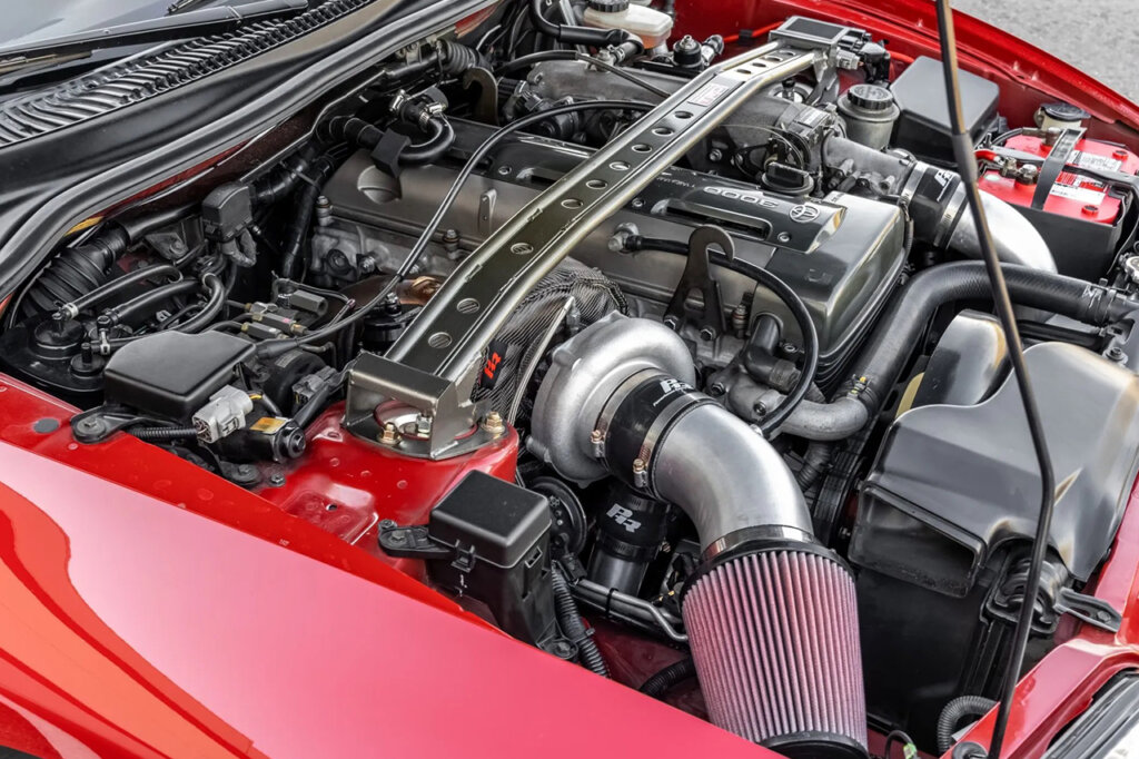 2JZ engine with a strut bar going over the top of it and the engine is in a red Toyota Supra