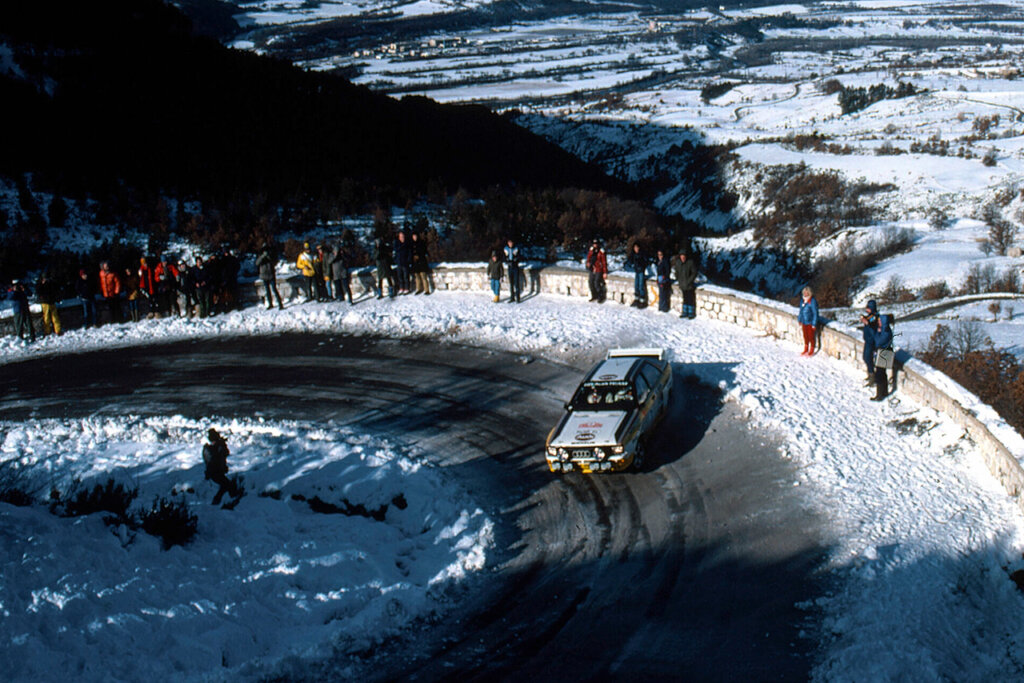 Audi rally car drifting through a snowy mountain turn, people standing off to the sides and overlooking a snow covered valley