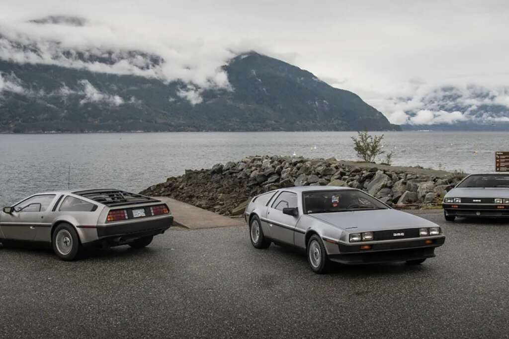 3 silver cars parked next to a boat launch dock. Cloudy sky and mountains behind them and a large lake