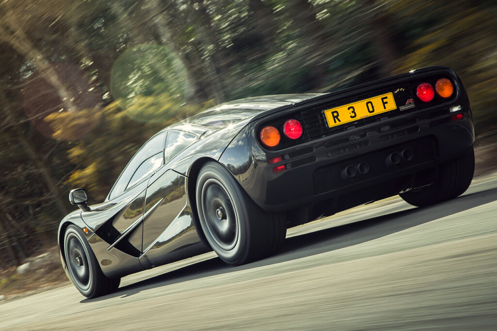 Grey McLaren F1 driving fast passing trees on road, red taillights and yellow license plate