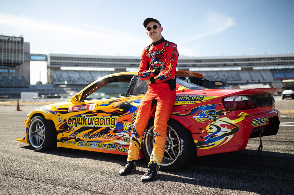 Man standing next to a yellow and orange Nissan Silvia S15. Sun shining and clear sky in background