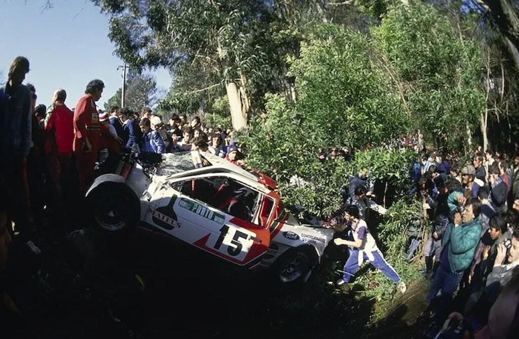 White Ford RS200 crashed into a ditch of spectators with trees in background and people pushing the car out