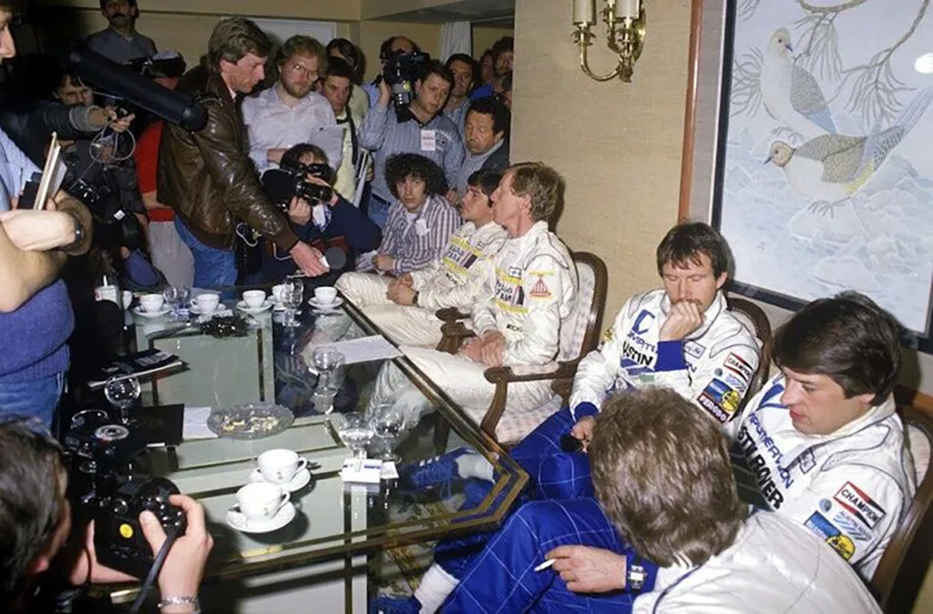 Group of racecar drivers in a hotel room speaking amongst each other and cameras