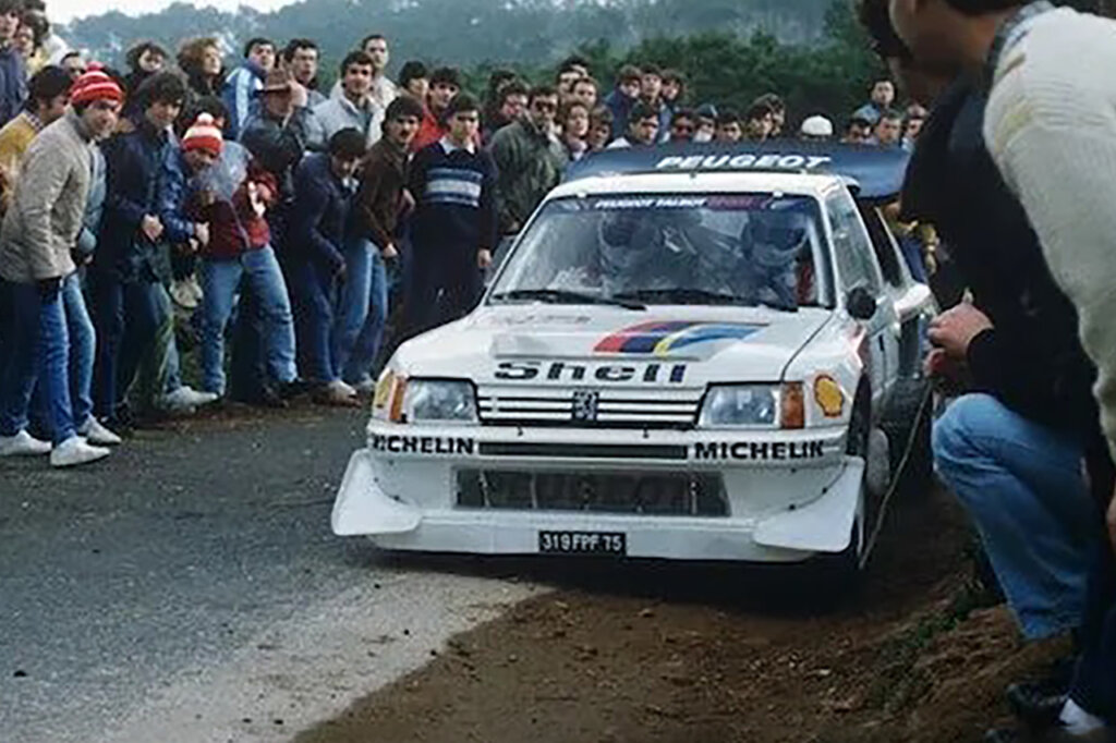 white Peugeot Group B rally race car inches away from spectators on the side of the road