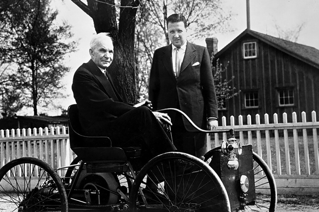 Two men in suits one sitting in a quadcycle and the other behind it.
