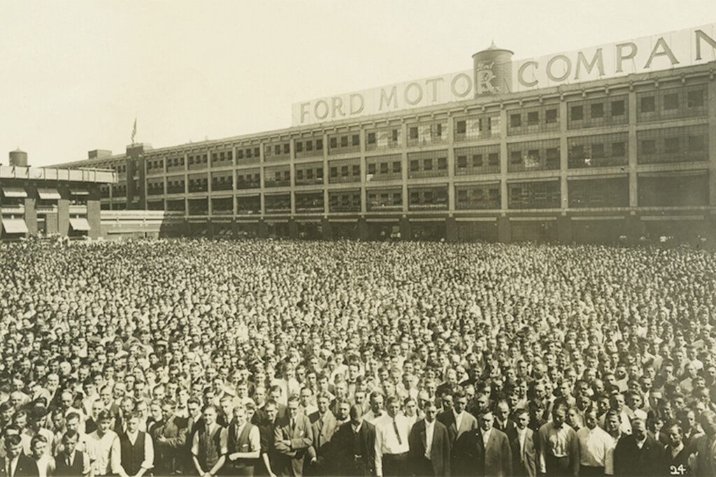 grey photo of a crowd of people standing outside of a large building that has a sign that says Ford Motor Company