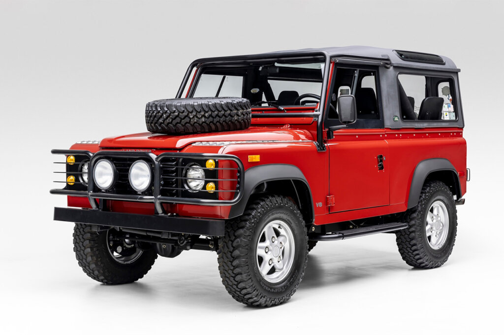 Red Land Rover 90 with a tire on the hood parked in a white room