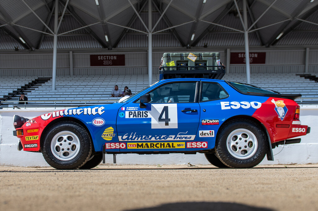 Blue and red Porsche 944 safari car parked on sand near stadium seats
