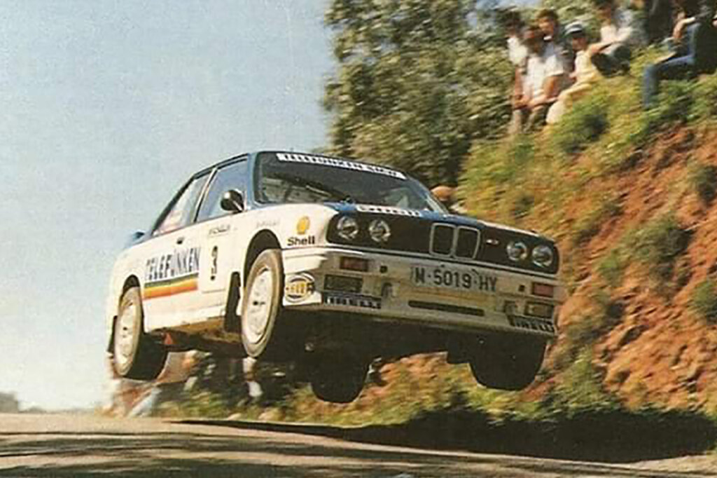 White BMW E30 M3 flying through a jump passing a mountainside