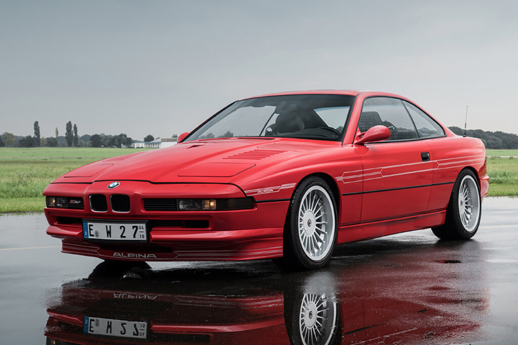 Red BMW Alpina B12 8-Series parked on wet ground showing its reflection with green grass in background