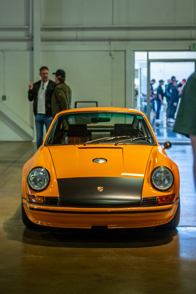 Orange Porsche 911 carrera with an orange strip over the hood with people behind it