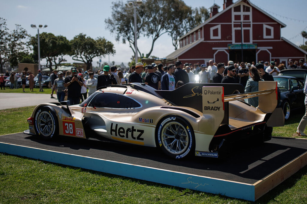 White and brown porsche car in front of a crowd of people with a red barn in the background