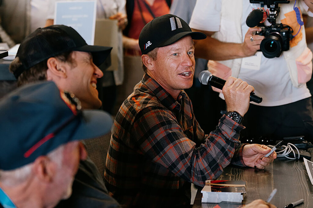 three men with hats on one a holding a microphone in speaking in front of a crowd of people