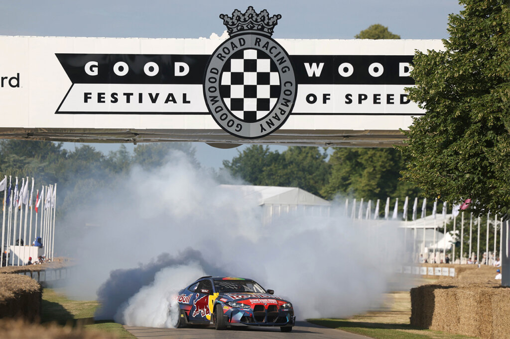 BMW race car drifting and leaving a trail of smoke behind it passing a black and white sign that says Goodwood Festival of Speed