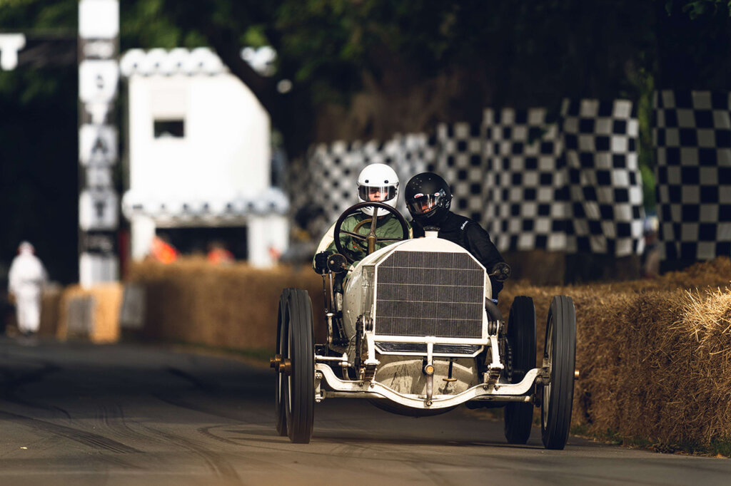 Two men in race gear driving an old motor vehicle on a road