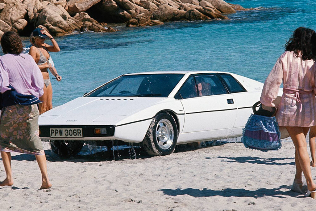 White Lotus espirit with a man in the drivers seat on a beach surrounded by people