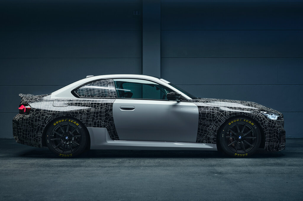 Side profile of a silver BMW with black graphics on the front and back ends of the car with a solid grey wall behind the car