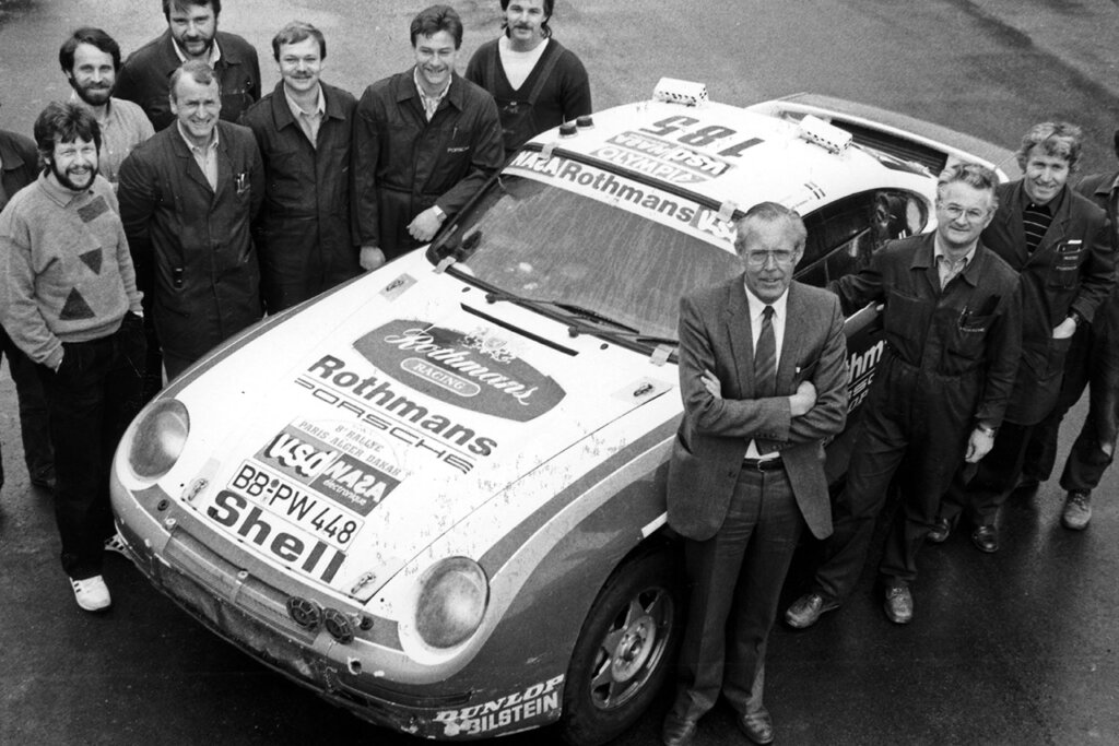 Black and white photo of several men in suits standing next to a Porsche race car