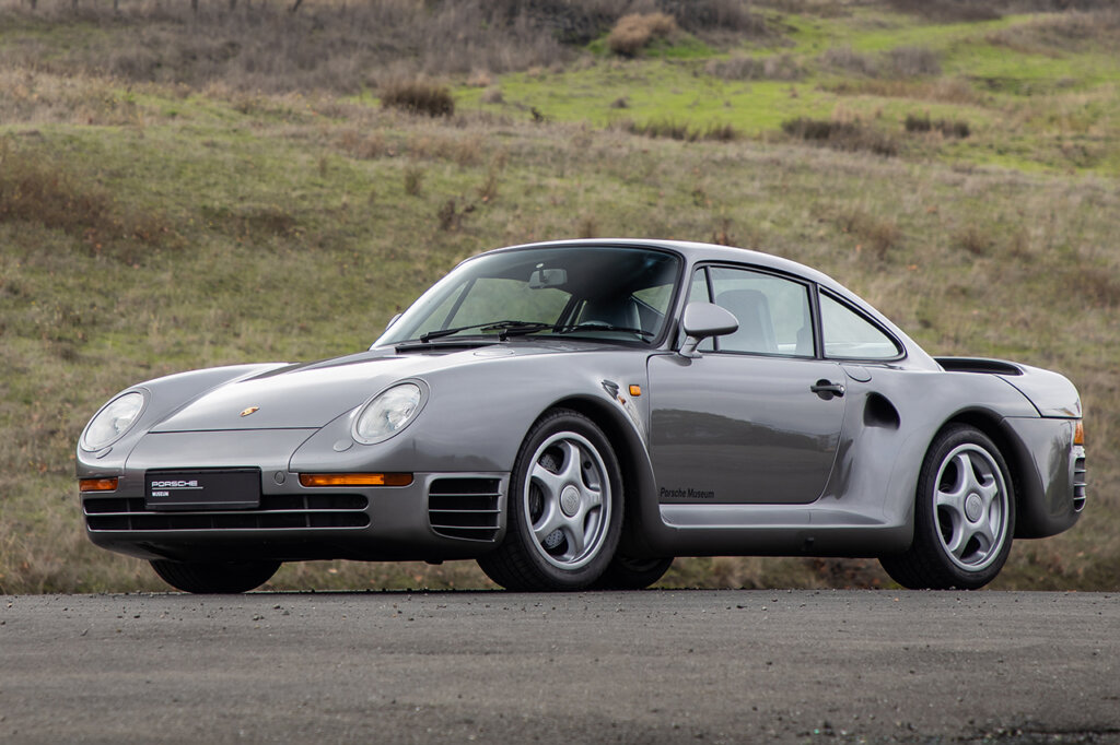 Grey Porsche 959 sportscar parked next to a grassy hill in background