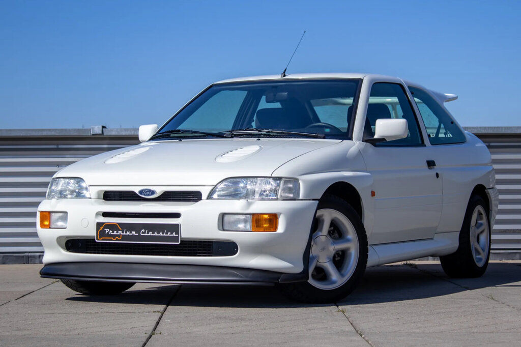 White Ford Escort Cosworth parked on a road with a fence behind the car and clear blue sky in background