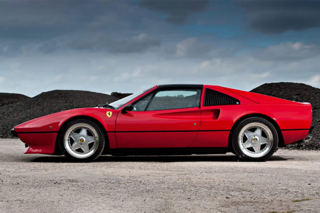side profile of red Ferrari parked on dirt ground with dark clouds in background