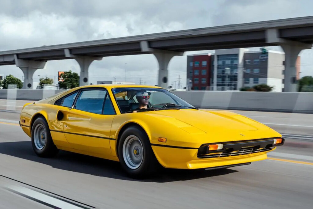Yellow Ferrari 308 driving fast on a freeway passing structures in background