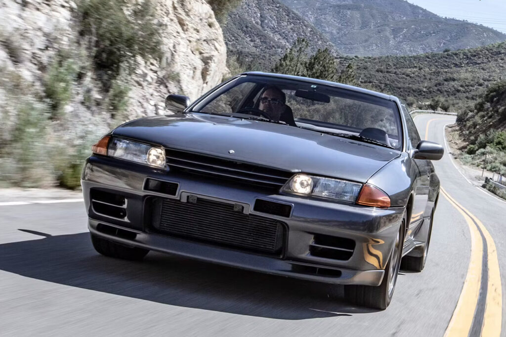 Grey Nissan R32 driving on a highway road with its lights on