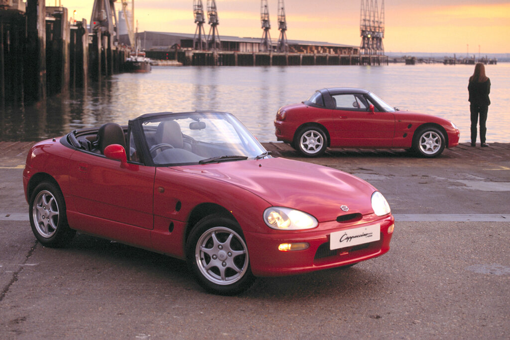 Two red cars parked next to each other one with headlights on and the other next to a body of water and a figure standing by the front end