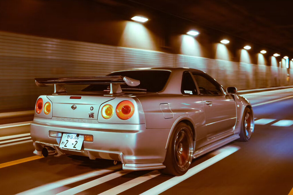 Silver R34 Skyline driving through a tunnel with several lights in the background and white solid lines on the roadway