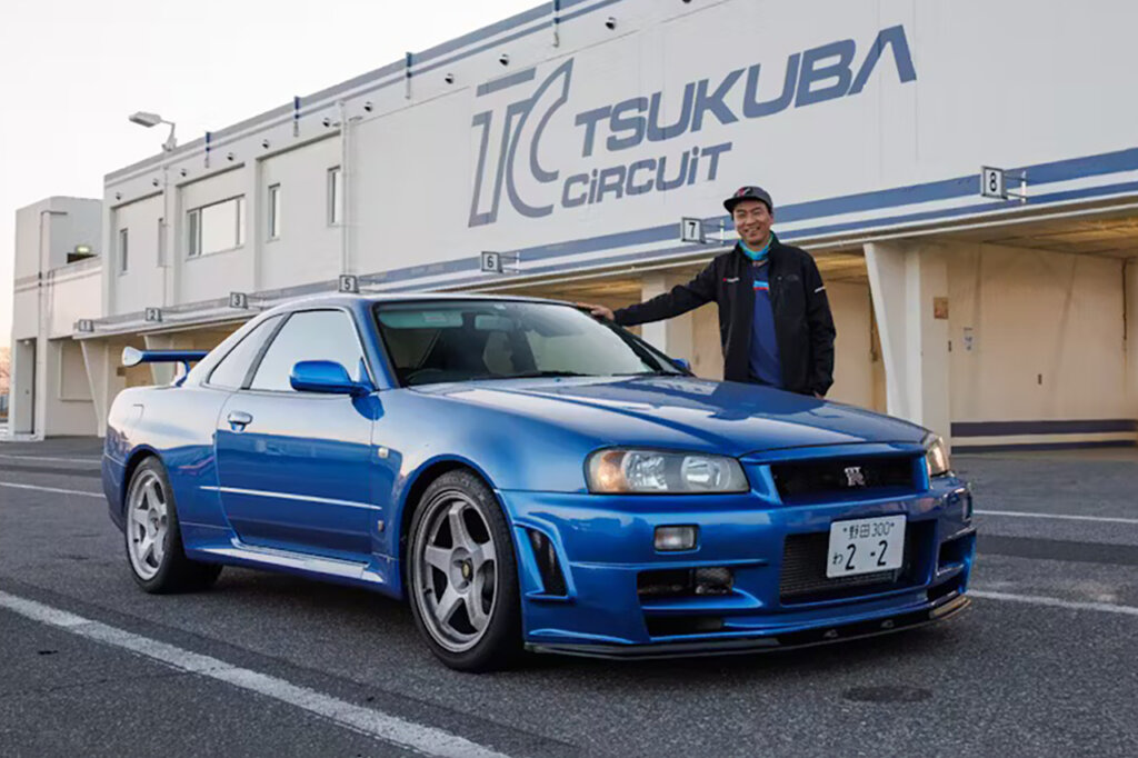 Blue Nissan Skyline GT-R R34 parked in front of a building that has a large sign on it with a man standing next to the car on the passenger side