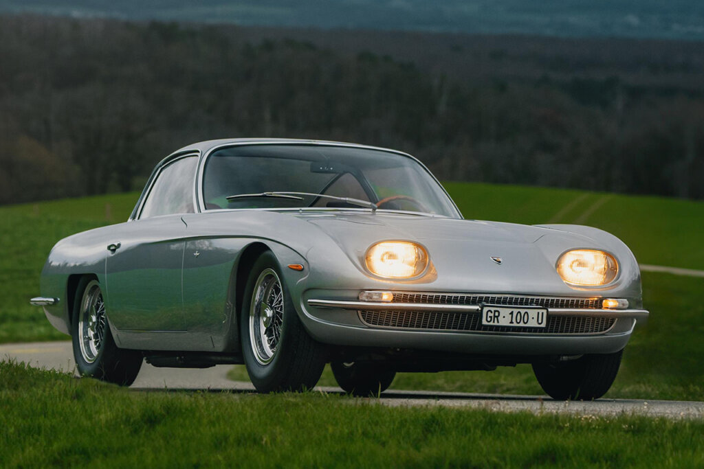 Silver Lamborghini 350 GT with headlights on parked on a road with grass in front and behind the car with trees in the background