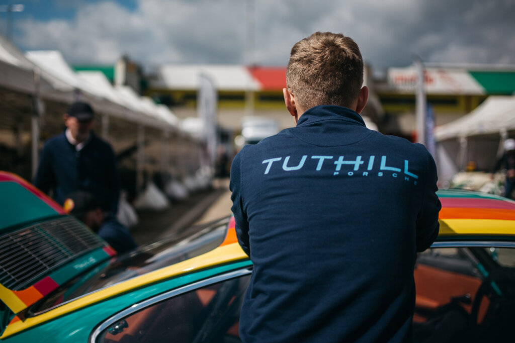 Man wearing a blue jacket that has the word Tuthill spelled out in light blue, man is leaning on a rainbow colored car facing away from camera