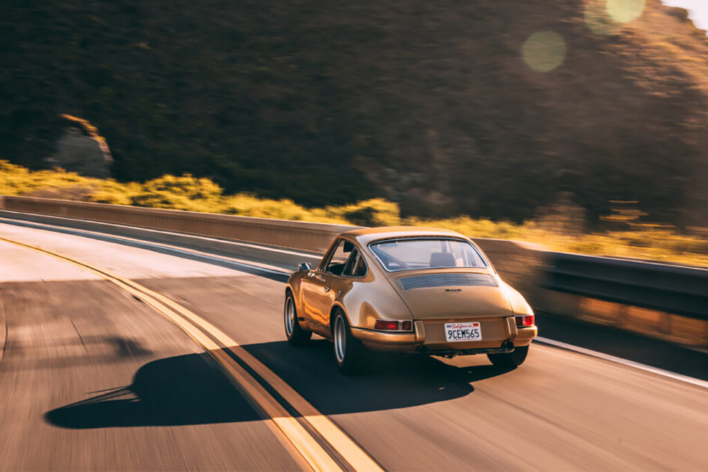 Gold Porsche 911 following a road going left with the sun peaking through the hills