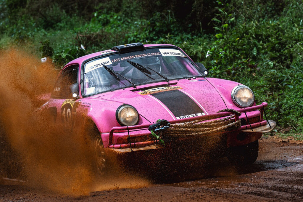 Pink Porsche car driving through mud and leaving behind a spray of mud passing dark green foliage