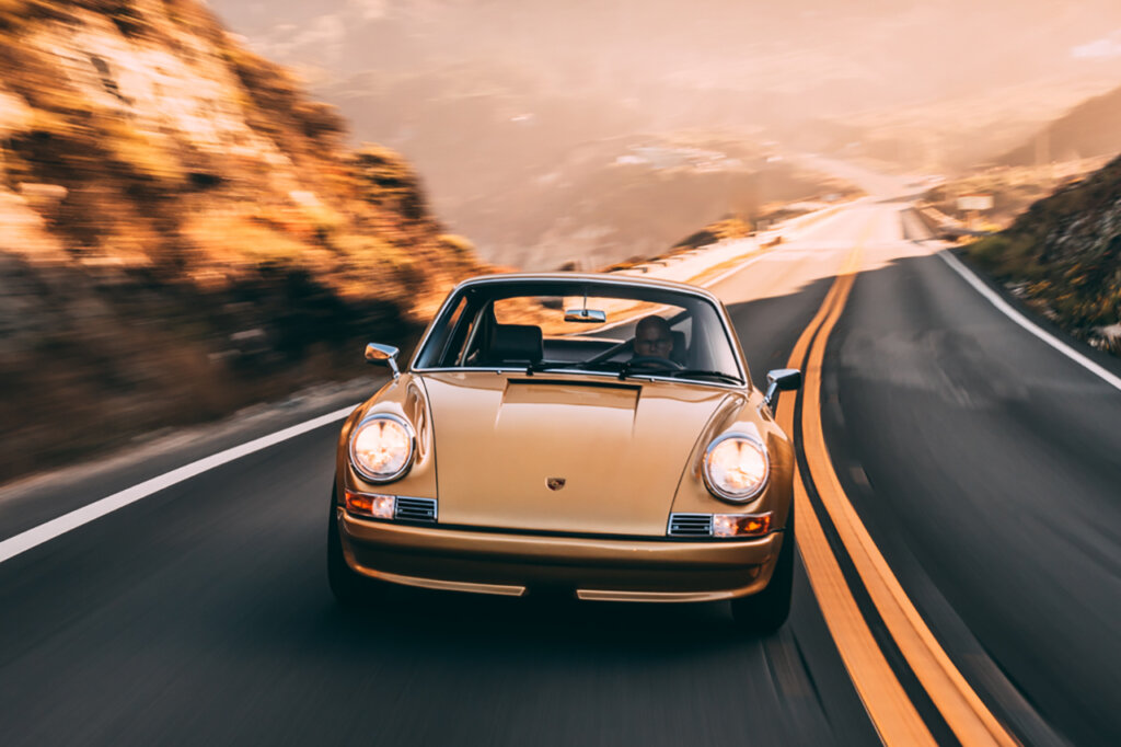 Man behind the wheel of a gold Porsche passing a hill on the left side of double yellow lines on the road