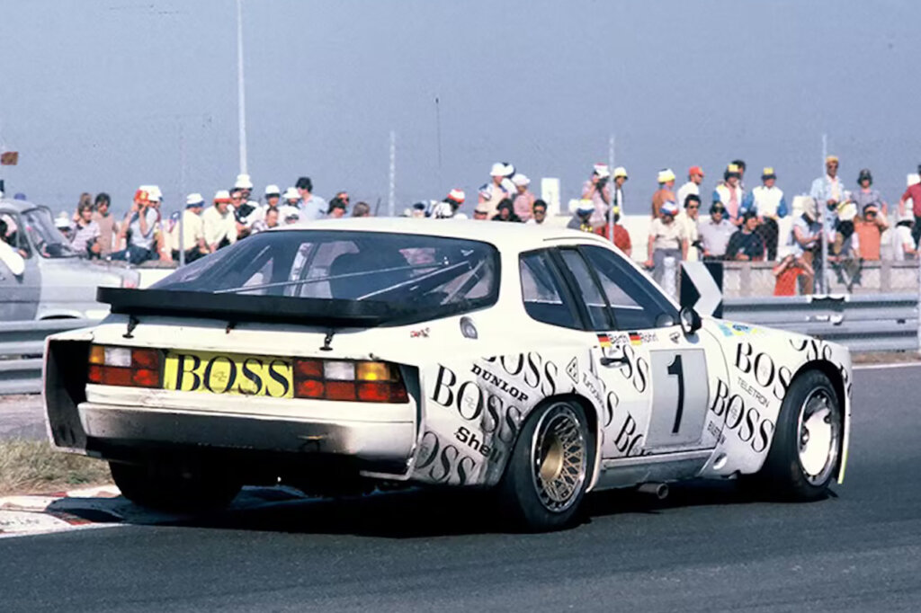 White Porsche 944 car with the word BOSS all over it at an angle driving on a road with people standing on the sides