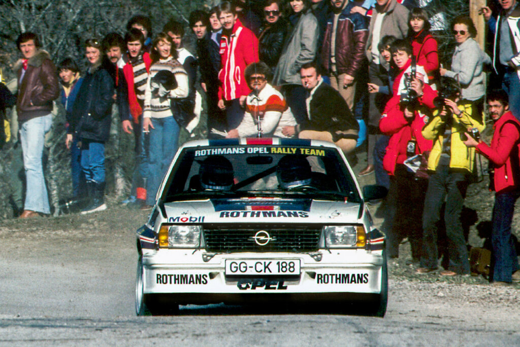 White Opel rally car driven by two people inside of the car passing a crowd of spectators on the side and behind the car