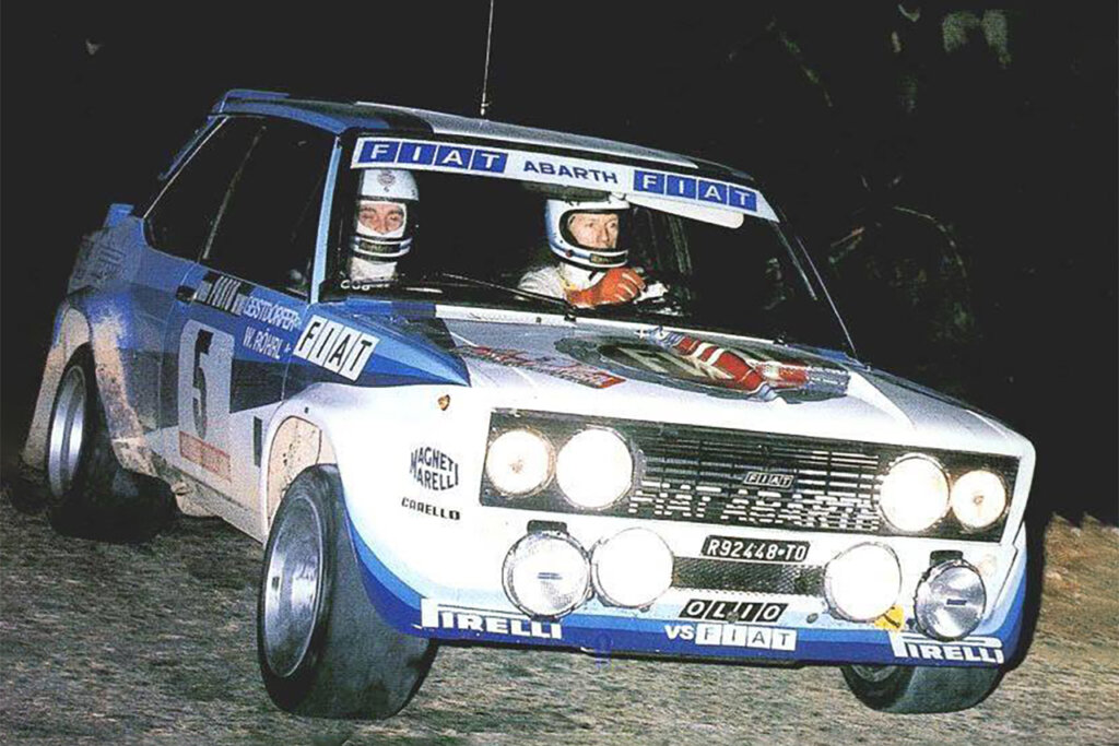 White and blue Fiat with headlights on and two race car drivers wearing helmets and gloves inside the car driving during the night