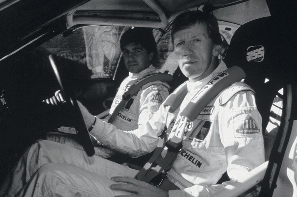 Black and white photo of two men in race suits looking at the camera sitting inside of a car