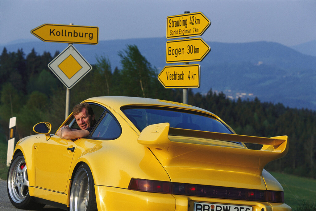 Man sticking his head out of the driver window of a yellow Porsche 911 parked on the side of a road with yellow signs pointing in several directions with words on it and trees and mountains in background