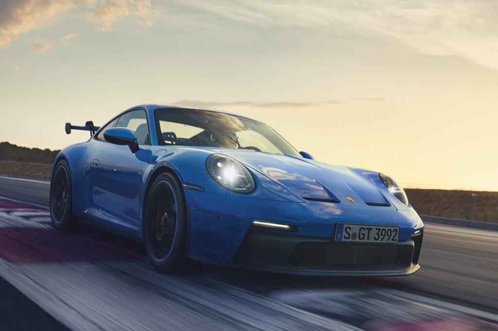 Blue 911 992 driving on a race track through a turn with the headlights on and the sun setting in the background