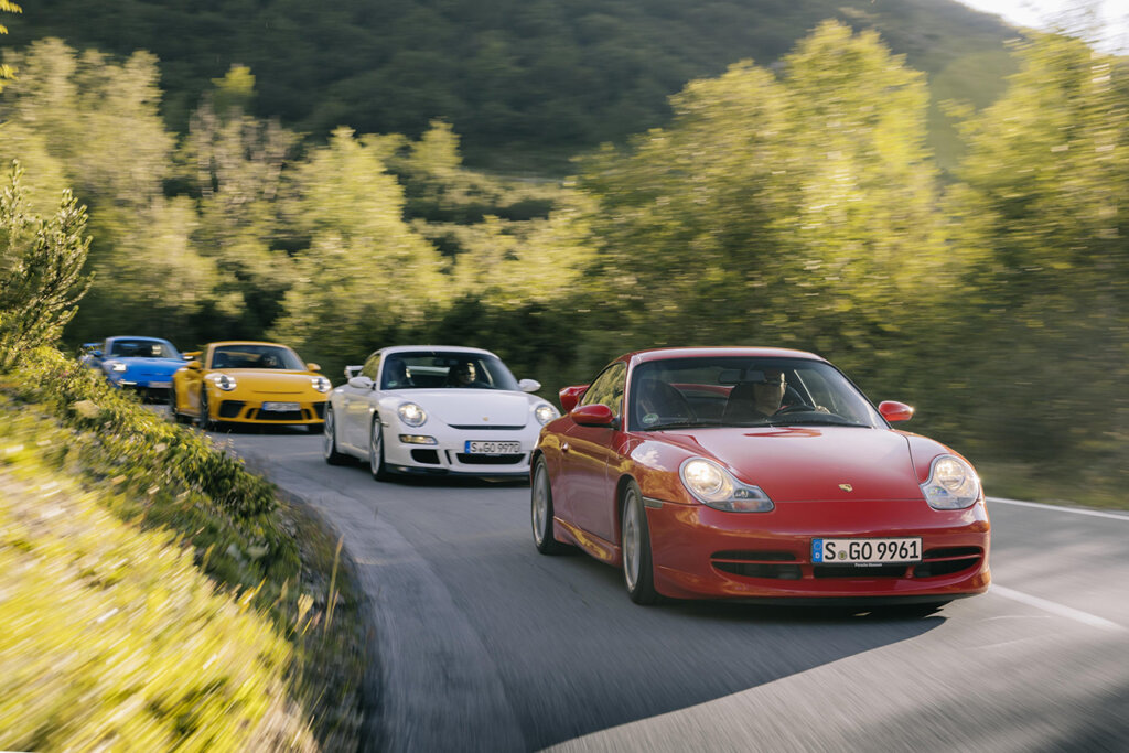 4 Porsche 911's driving closely in a line, lead car is red, then white, then yellow and a blue one at the end with trees on the sides of the cars