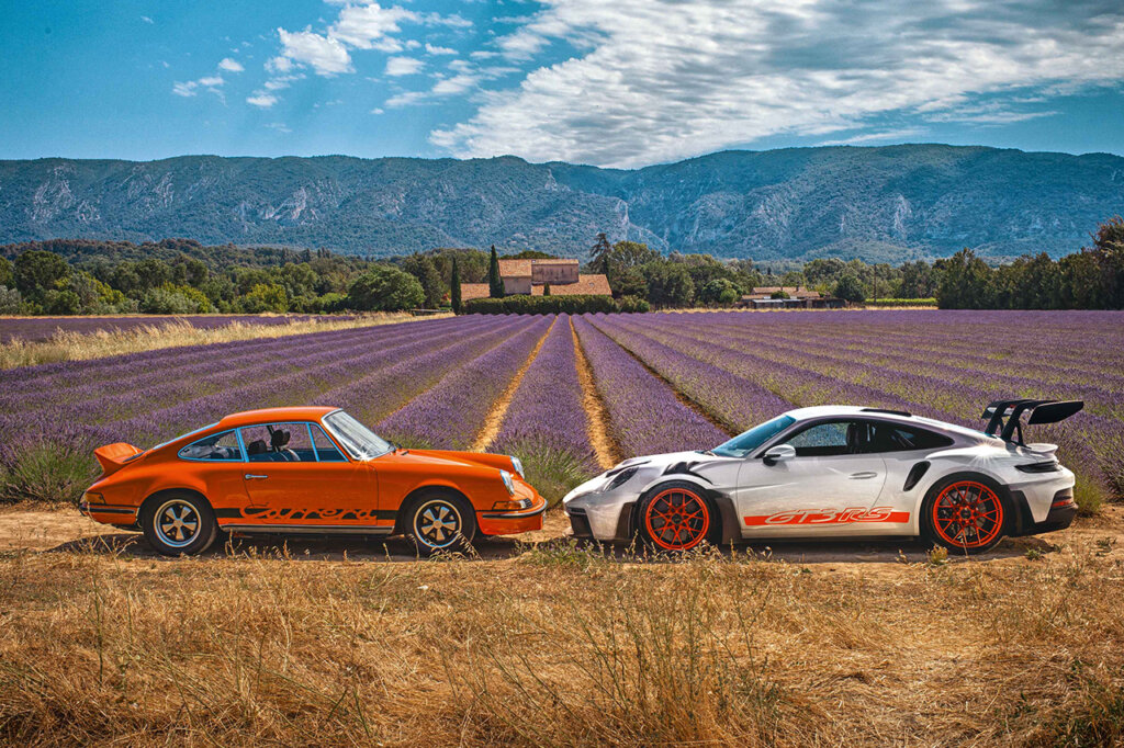 two Porsches parked in front of eachother with noses close to touching one another with a field of purple flowers in the background leading to a house and hills in the distance