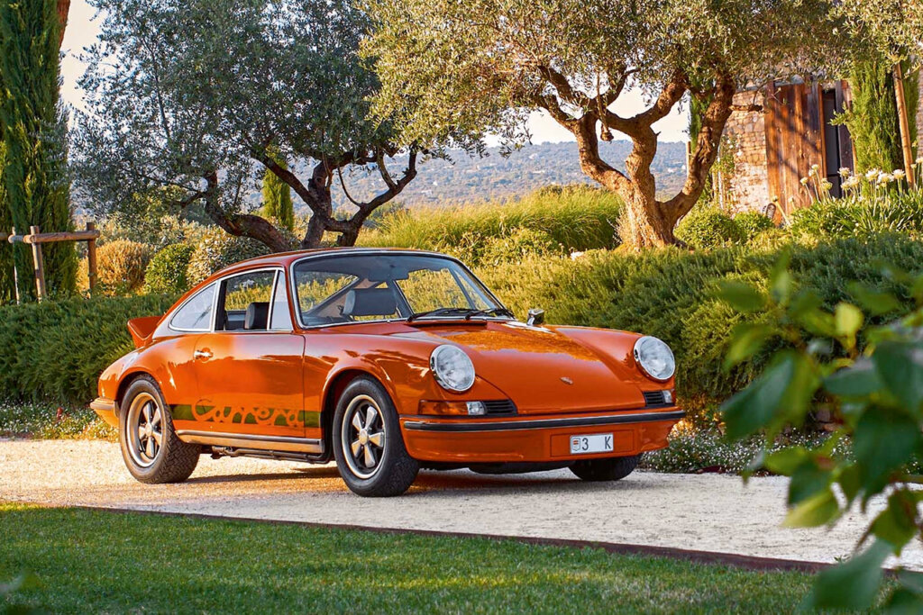 Orange 911 Carrera RS parked in a driveway with flowers, trees and hills in the background