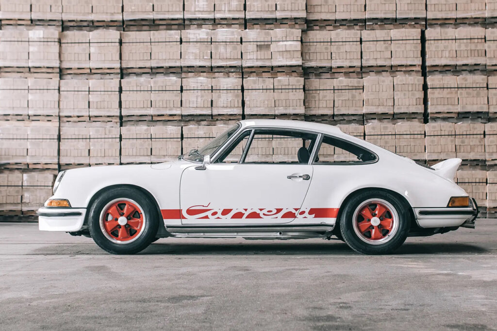 White Porsche 911 with red decals that says Carrera on the side of the car with red rims parked in a garage with stacks of boxes behind the car