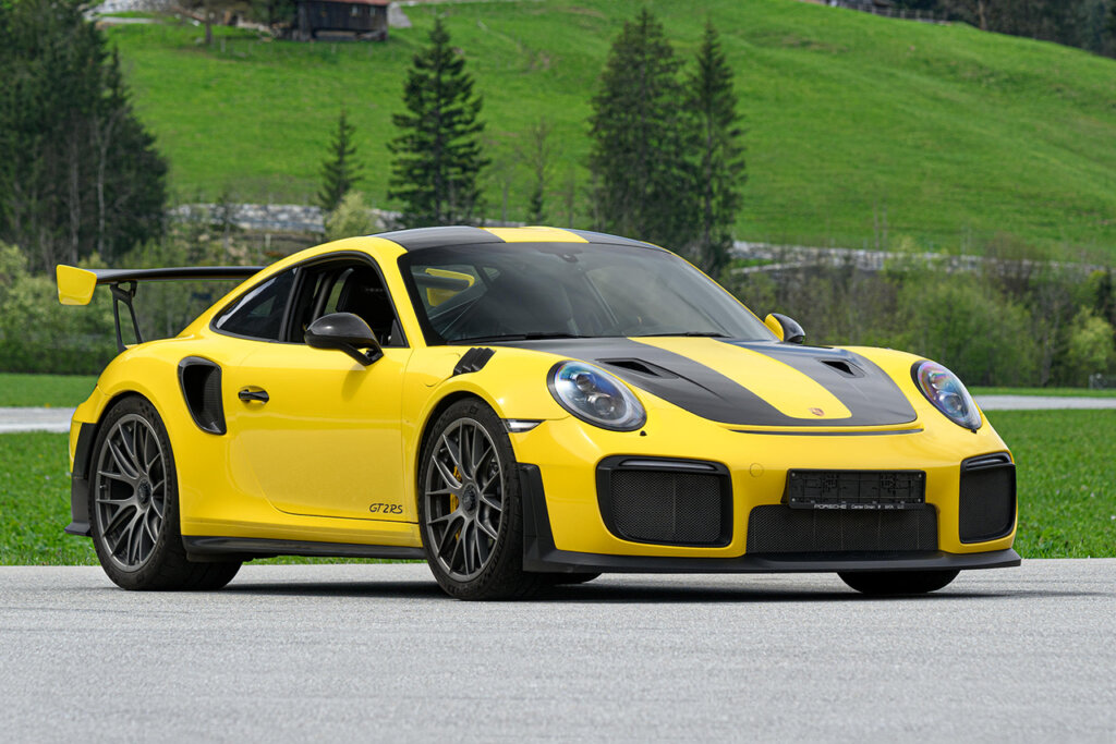 Yellow and black 991 GT2 RS with large wing and aerodynamic bumper vents parked in front of trees and large green hills behind it