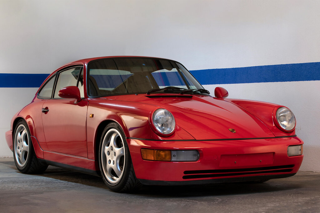 Red 911 964 RS parked near two walls joined together with a solid blue stripe going behind the car and continuing to the other side 